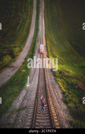 Aerial view A male hipster in a cap holding up a hand with a gesture Victory is standing on rails in front of an approaching tra Stock Photo