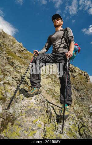 Porter of a stylish hipster traveler with a beard and a backpack in sunglasses and a cap with trekking poles standing on a rock Stock Photo