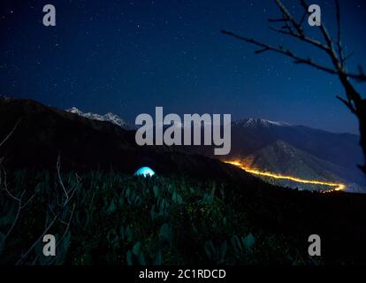 Blue gloving tent at camping in the mountain valley under night sky with stars and city light at background Stock Photo