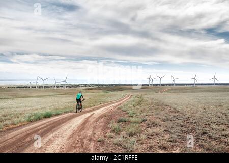 Man on mountain bike rides on the road to the wind turbines farm near the lake Against blue cloudy sky. Ecological and zero waste lifestyle concepts. Stock Photo