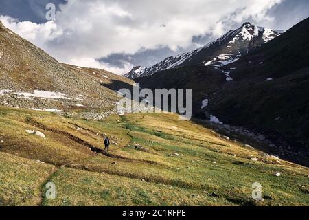 Small tourist with backpack is walking in the green mountain valley with snowy peaks and cloudy sky background Stock Photo