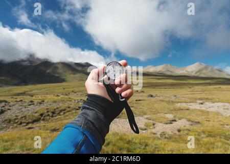 Viewpoint shot. A first-person view of a man's hand holds a compass against the background of an epic landscape with cliffs hill Stock Photo