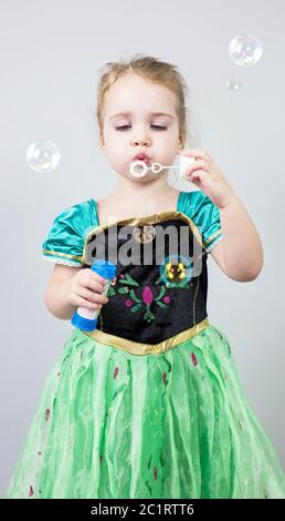Beautiful little blonde girl, has happy fun cheerful smiling face, pink dress, soap bubble blower. Portrait with pink and white Stock Photo