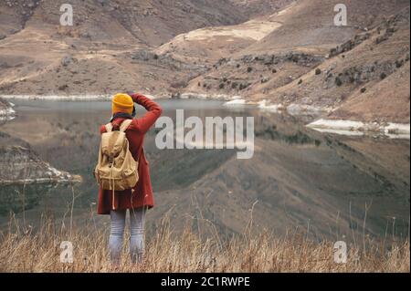 A portrait from the back of a girl traveler take a picture on the background of a lake in the mountains in autumn or early sprin