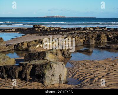 Rocky and sandy shore at Bamburgh, Northumberland, England, with a view to Inner Farne Lighthouse in the Farne Islands Stock Photo