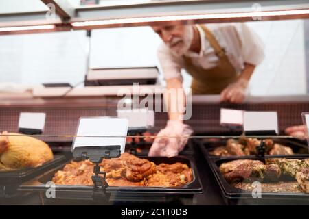 Close up of raw meat pieces in refrigerator with price tags ready for sale in meat department. Senior male butcher putting out of glass counter plate with sliced fresh steaks. Concept of food. Stock Photo