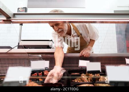 Close up of raw meat pieces in refrigerator with price tags ready for sale in meat department. Senior male butcher putting out of glass counter plate with sliced fresh steaks. Concept of food. Stock Photo