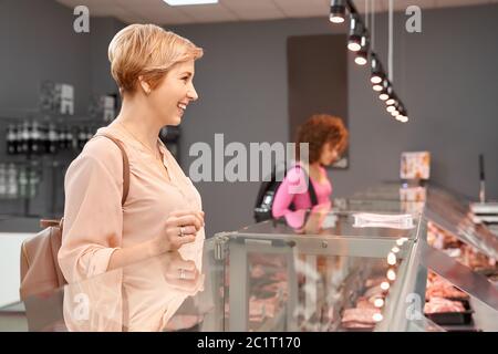 Side view of fasionable smiling ladies looking and choosing fresh meat in store. Cheerful women standing near store counter with big assortment of raw meat products in supermarket. Stock Photo