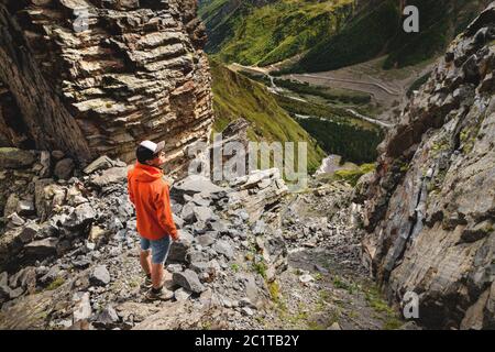 Portrait of a male hiker in shorts and a cap from the back is standing on high rocks at the edge of a cliff against the backgrou Stock Photo