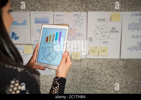 Unrecognizable woman standing in front of papers on wall holding digital tablet computer working with data stats and graphs Stock Photo