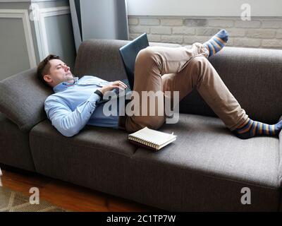 A man lies at home on the couch and works on a laptop. Remote work concept. Stock Photo