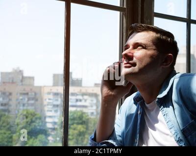 Portrait of a young man who stands by the window and makes a phone call. Communication concept. Stock Photo
