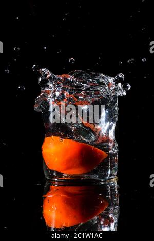 A glass of water in a dark background and water being splashed with a piece of cut tomato dropped inside the glass Stock Photo