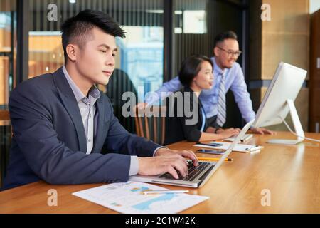 Handsome young man sitting at office table searching something on his laptop while his colleagues working together Stock Photo