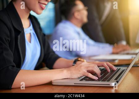 Unrecognizable woman sitting at table in office with her colleagues typing something on her laptop Stock Photo