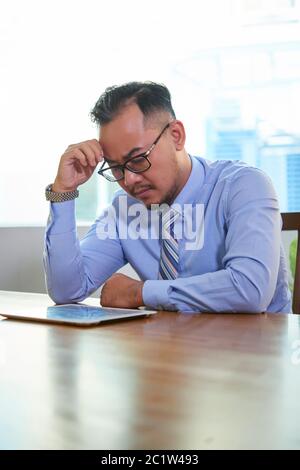 Pensive Asian man wearing eyeglasses sitting at office desk with reading important documents using digital tablet Stock Photo