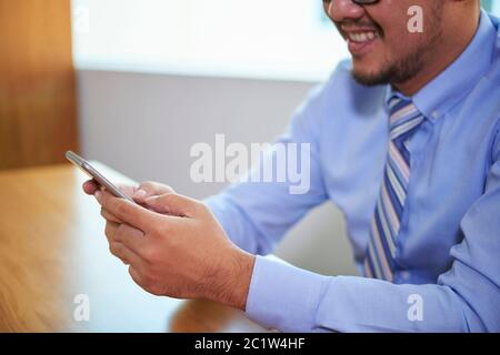 Cheerful man wearing light blue shirt and tie sitting at office desk using his smart phone at work, horizontal shot Stock Photo