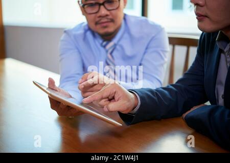 Two Asian male office workers sitting together at table using Internet on digital tablet, horizontal shot Stock Photo