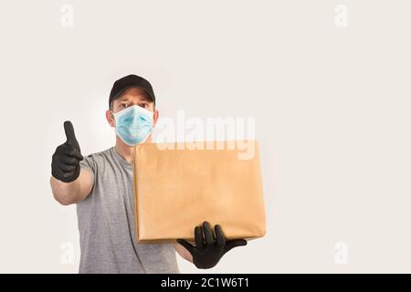 Delivery man with medical gloves and mask holding a box and with thumb up in a front view Stock Photo