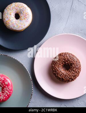 Three donuts on plates, chocolate, pink and vanilla donut with sprinkles, sweet glazed dessert food on concrete textured background, top view Stock Photo
