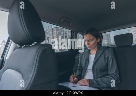 Businesswoman doing business paperwork on car back seat, adult caucasian female businessperson executive analyzing business results while traveling by Stock Photo