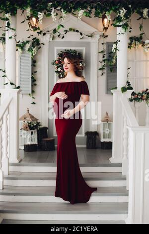 Charming pregnant woman in marsala dress posing on steps of house. Stock Photo