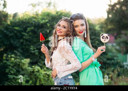 Two pretty and fashionable girlfriends holding candies on stick. Stock Photo