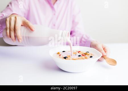 Close up of female pouring fruit yogurt into bowl with cereals, strawberries and blueberries. Stock Photo