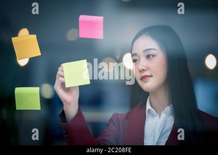 Portrait of Young asian businesswoman writing sticky paper pad on glass in modern office. Business plan concept. Stock Photo