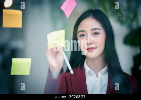 Portrait of Young asian businesswoman writing sticky paper pad on glass in modern office. Business plan concept. Stock Photo
