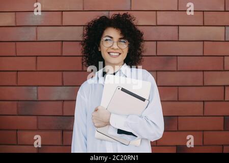 Curly haired businesswoman with eyeglasses smiling at camera on a brick wall outside with a computer Stock Photo