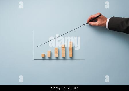 Hand of a businessman drawing a growing arrow above a column chart made of wooden blocks. Stock Photo