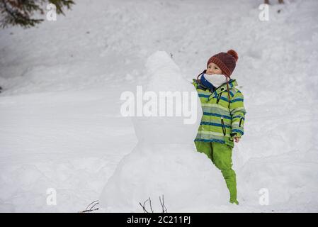 Toddler child in green winter suit and red hat making a snowman. Stock Photo