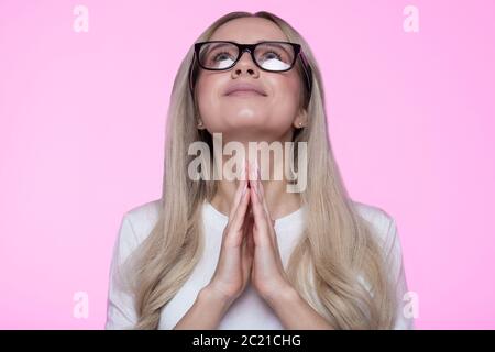 Closeup portrait of young glad smiling female in eyeglasses keeps palm together, looking up, hopes for the best in life, prays to have good luck. Peac Stock Photo