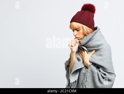 Cute sick young teen woman in red hat, wrapped in warm scarf coughing, closed eyes.Female feeling the first symptoms of illness, isolated.Bronchitis, Stock Photo