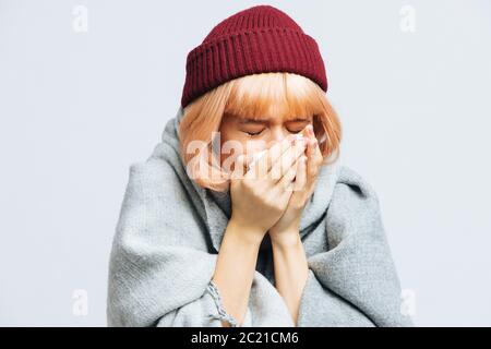 Unhealthy young woman in red hat, warm scarf with paper napkin sneezing, experiences allergy symptoms, caught a cold, closed eyes. Sick desperate woma Stock Photo