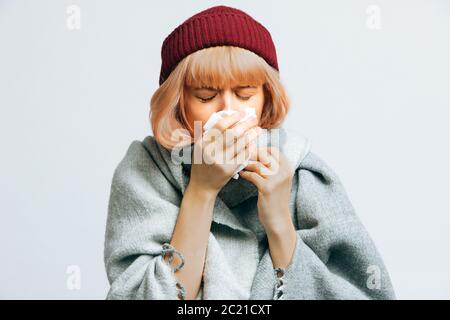 Unhealthy young woman in red hat, warm scarf with paper napkin sneezing, experiences allergy symptoms, caught a cold, closed eyes. Sick desperate woma Stock Photo