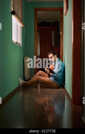 Young man plays guitar sitting in the hallway of his house Stock Photo