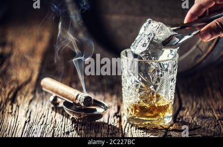 Ice cube being put into an ornamental cup of whisky placed on a old fashioned wood and burning cigar aside Stock Photo