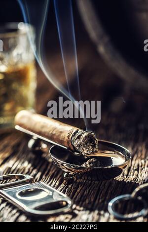 Burning cigar with cutter and a glass of whiskey in the back with blueish smoke rising Stock Photo