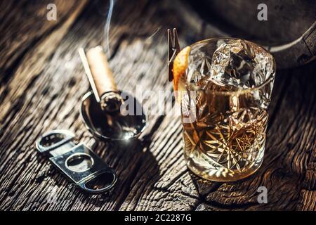 Ornamental glass full of whiskey and ice on a vintage wood next to a cigar cutter and a burning cigar Stock Photo