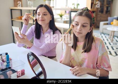 Pretty mother and teenage daughter sitting at table in living room and using brushes while putting on makeup