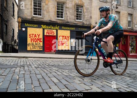 Edinburgh, Scotland, UK. 16 June, 2020. As shops open in England, Scottish shops and businesses remain closed, Streets are empty and pubs and shops are still closed with many boarded up. Bars might be allowed to open outside areas at end of week but currently they are only-permitted  to serve drinks to takeaway. Pictured; Cyclist rides past boarded up restaurant on Victoria Street.   Iain Masterton/Alamy Live News Stock Photo