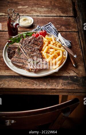 Barbecued or grilled T-bone steak and French fries served with salad trimming on an old rustic table in a tavern for lunch Stock Photo