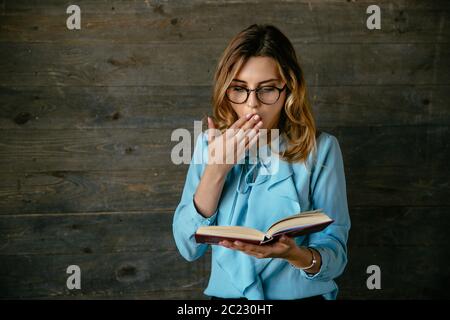 Young pretty woman in eyeglasses looks shocked, cover her open mouth with hand while reading a book. Dressed in elegant blouse. Stock Photo