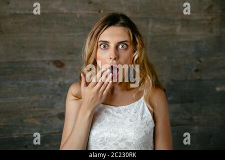 Pretty young woman surprised about something, covering her open mouth by hand, with widely eyes. Dressed in white singlet. Stock Photo