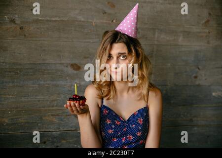 Attractive girl with frustrated face after blowing out the candle on the cupcake. Wearing festive hat, curly hair. On grey wood background. Stock Photo