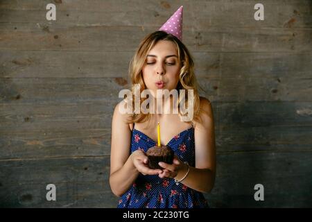 Cute young woman blowing out candle on birthday chocolate cupcake. Festive hat, curly hair. Holidays concept. Stock Photo