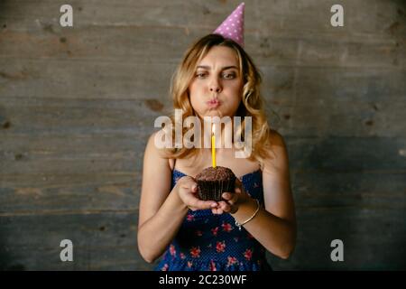 Beautiful young woman blowing out candle on birthday cupcake. Festive hat, curly hair. Holidays concept. Stock Photo