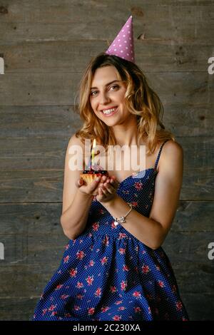 Charming smiling young woman with curly hair, celebrating own birthday, holds a fruit cupcake in hands. Wearing colorful dress, festive hat. Holiday c Stock Photo